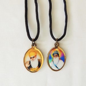 May include: Two oval pendants featuring religious figures, each suspended from a black cord. The pendants have gold-colored frames and detailed portraits. One figure wears a yellow turban, the other a blue and white turban.
