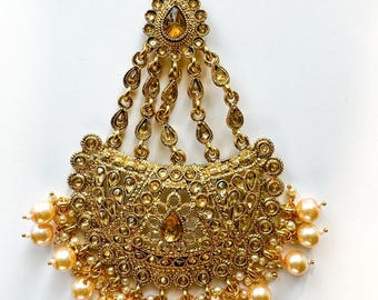 May include: Gold-toned headpiece with intricate filigree and teardrop accents. The ornate design features a central amber-colored stone and is adorned with dangling chains and faux pearls. A hook is at the top for securing the headpiece.