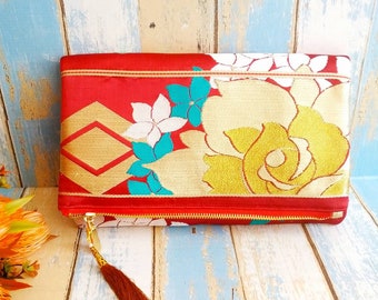 May include: A red clutch purse with a gold zipper and a brown tassel. The purse is decorated with a floral pattern in shades of white, yellow, and turquoise.