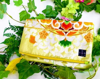 May include: A yellow clutch purse with a floral pattern and a yellow tassel. The purse has a zipper closure and a small black label with the text "nota" on it.