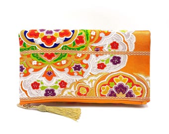 May include: A bright orange clutch purse with a gold tassel. The purse is decorated with a colorful floral pattern in shades of red, orange, yellow, green, purple, and white.
