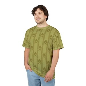 May include: A short-sleeved, olive green t-shirt with a repeating arrow pattern. The shirt is made of a soft material and is worn with blue jeans. The design features vertical stripes of arrows in varying shades of green.