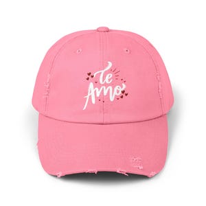 May include: Pink distressed baseball cap with the phrase "Te Amo" in white script, accented with small red hearts. The cap has a curved brim and a distressed, worn look.