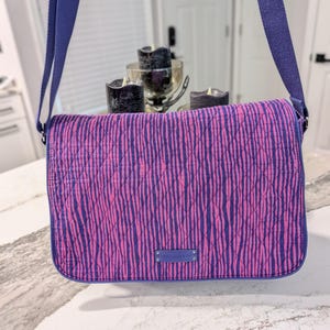 May include: A quilted messenger bag with a vibrant pink and purple vertical stripe pattern. The bag has a blue adjustable shoulder strap and trim. A small blue rectangular tag is centered on the front.