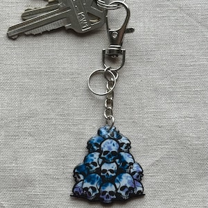 May include: A keychain with a silver clasp and chain, holding a cluster of blue and purple skull-shaped charms. The keychain is attached to a set of silver keys. The skulls have a watercolor effect.