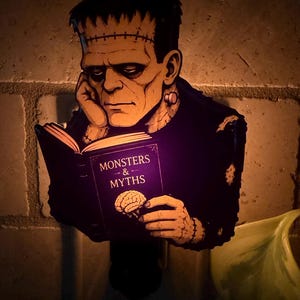 May include: A plug-in night light featuring a graphic illustration of Frankenstein's monster reading a purple book titled Monsters & Myths with a brain icon on the cover. The monster has green skin, a flat head with stitches, and a dark shirt.