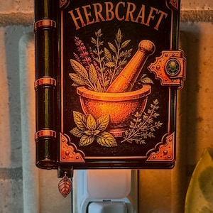May include: A plug-in night light shaped like a book with a black cover and copper-coloured accents. The cover features the text HERBCRAFT above an illustration of a mortar and pestle filled with various herbs. A small leaf charm hangs from the spine.