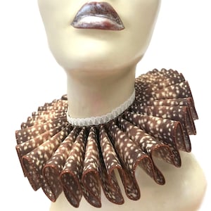 May include: A brown and white patterned ruffled collar with a white lace trim. The collar is made of fabric and is worn around the neck.