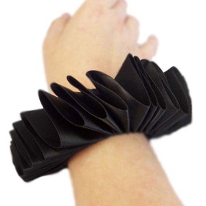 May include: A black satin ribbon wrist cuff with multiple pleats. The cuff is worn on a person's wrist.