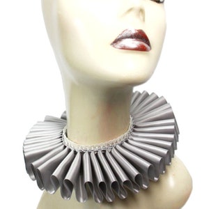May include: A silver ruffled collar made of satin. The collar is worn by a mannequin with a neutral expression. The collar is a popular accessory for historical reenactments and costume parties.