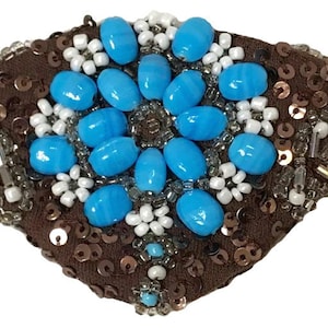 May include: A brown eye patch with a blue and white beaded design. The patch is made of fabric and has a sequined border.