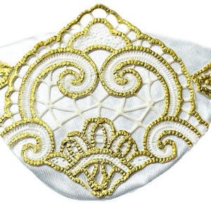Pode incluir: Uma máscara de olhos de cetim branco com acabamento em renda dourada e uma faixa elástica dourada. A máscara apresenta um design decorativo de renda dourada no centro, com um padrão geométrico e detalhes em espiral.