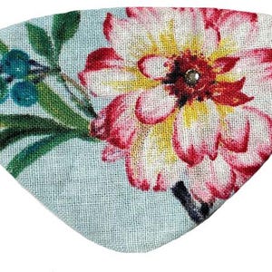 May include: A floral eye mask featuring a large flower with red and pink petals, yellow center, and a small jewel. The mask has a light blue background with green leaves and berries. A black elastic band is attached.