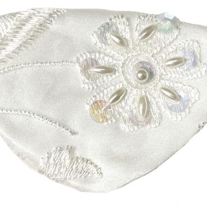 May include: A white satin eye patch featuring white floral embroidery, iridescent sequins, and small white faux pearls arranged in a flower pattern. The patch is attached to a tan elastic strap extending from both sides of the fabric.