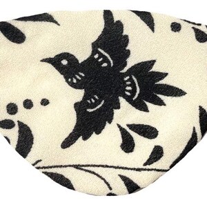 May include: Cream-colored eye mask with a black bird and floral pattern. The mask has an elastic band for a secure fit. The bird is in flight, surrounded by leaves and dots.