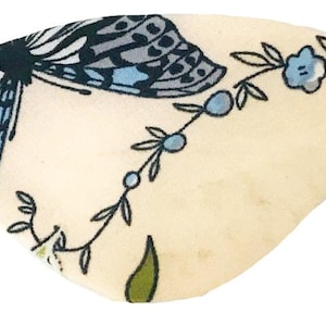 Può includere: Un paraluce bianco con un motivo a farfalla blu e floreale. La farfalla ha ali nere e grigie con accenti blu. I fiori sono blu e bianchi.