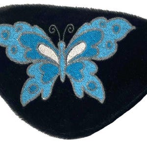 Pode incluir: Tampão de olho preto com um desenho de borboleta azul. A borboleta tem asas azuis com contornos prateados e um corpo branco. O tampão de olho tem uma faixa elástica para fixá-lo.