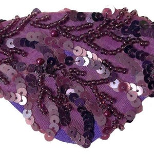 Pode incluir: Um tapa-olho roxo com um arranjo denso de lantejoulas roxo-escuras e pequenas contas dispostas num padrão orgânico ramificado. A base de tecido é de um roxo vibrante e o tapa-olho está preso a uma fina tira elástica preta em cada lado.