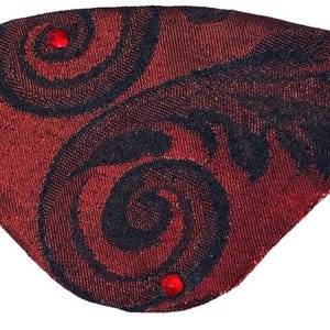 May include: Red and black patterned eye patch with a black elastic band. The eye patch features a swirling design and is embellished with two red gemstones. The fabric has a textured appearance.