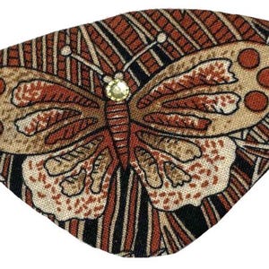 Pode incluir: Uma máscara de olhos decorativa com um desenho de borboleta. A borboleta tem asas bege com manchas castanhas e um corpo castanho, num fundo de riscas castanhas e pretas. Uma pequena joia brilhante adorna o centro da borboleta. A máscara tem um elástico preto.