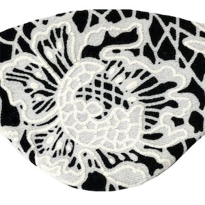 May include: An eye patch featuring a black base with a prominent white floral lace overlay pattern. The design displays intricate, interconnected petal and leaf shapes across the surface, secured by a thin black elastic strap on each side.