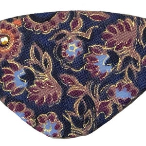 Könnte beinhalten: Ein blaues Augenpflaster mit einem floralen Muster in Violett-, Gold- und Orangetönen. Das Pflaster hat einen dekorativen Stein in der Mitte.