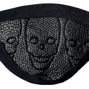 May include: Black eye patch featuring a textured surface with three embossed skulls. The eye patch is made of a dark fabric and has an elastic strap for securing it around the head. A gothic accessory.