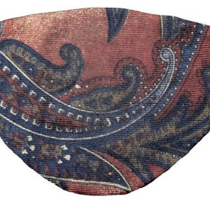 May include: A paisley patterned eye patch with a black elastic band. The eye patch features a design in shades of red, navy blue, and gold. The fabric has a textured appearance.