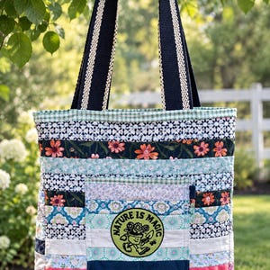 May include: A quilted tote bag featuring horizontal fabric strips in various floral and geometric patterns. A central square pocket displays a circular patch with a cartoon potted plant and the text "NATURE IS MAGIC". It has two dark blue straps.
