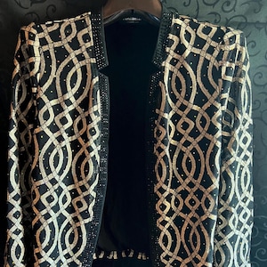 Puede incluir: Una chaqueta negra sin cierre con un intrincado patrón geométrico dorado. La chaqueta está adornada con pequeños acentos brillantes y tiene un forro negro. El diseño presenta una red de formas interconectadas.