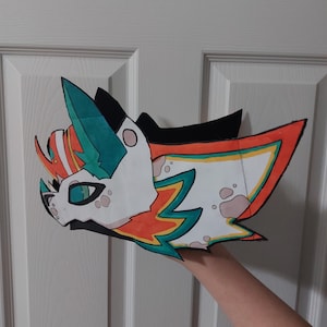May include: A colorful, stylized cartoon animal head with a white base, teal, orange, and black accents. The head has large, expressive eyes and is held up by a person's hand. The design is outlined in black.