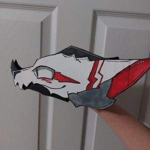 May include: A handmade, stylized mask in the shape of a wolf's head. The mask features a white base with red and gray accents, including lightning bolt designs. Black outlines define the features, and the mask is held up by a person's arm.