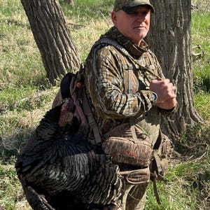 Puede incluir: Un cazador con ropa y sombrero de camuflaje lleva un gran pavo salvaje. El pavo tiene plumas oscuras y está colgado sobre el hombro del cazador. El cazador está de pie en una zona herbosa con árboles.