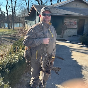 Puede incluir: Una persona con chaqueta y gorra de camuflaje, sosteniendo varios faisanes. La chaqueta tiene detalles amarillos. El fondo incluye un edificio y árboles, sugiriendo una escena de caza al aire libre.