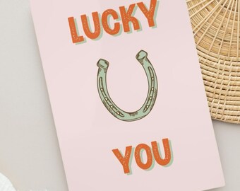 May include: A rectangular greeting card with a light pink background featuring the orange text LUCKY at the top and YOU at the bottom. Centered between the words is a line-drawn illustration of a light green horseshoe with dark outlines.
