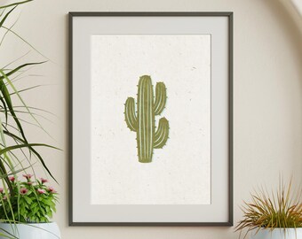 May include: A framed wall art print featuring a centered illustration of a green saguaro cactus with white vertical lines on a textured off-white background. The artwork is enclosed in a thin, dark-colored rectangular frame with a clean border.