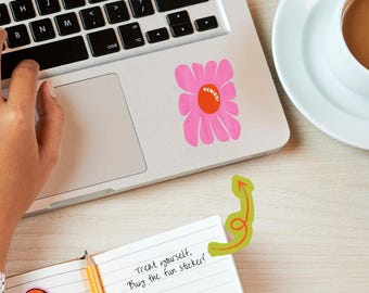 May include: A pink flower-shaped sticker with an orange center and the text HOWDY! in white, applied to a silver laptop. The laptop also features a small green arrow graphic pointing toward a notebook with handwritten text and several small circular stickers.
