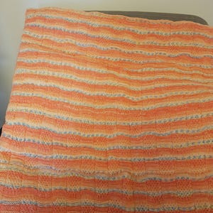 May include: A knitted blanket with horizontal stripes in shades of orange, peach, and cream, with subtle blue accents. The blanket is folded and appears to be made of soft yarn. The texture is visible, suggesting a handmade item.