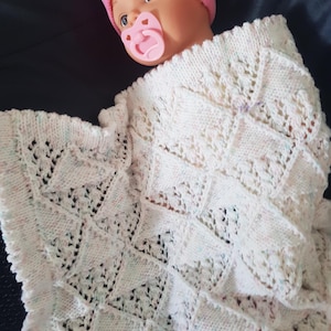 May include: A baby doll wearing a pink knitted hat and a dummy is wrapped in a white, hand-knitted blanket. The blanket has a geometric pattern with openwork details. The doll's face is visible, with blue eyes and a pink dummy.