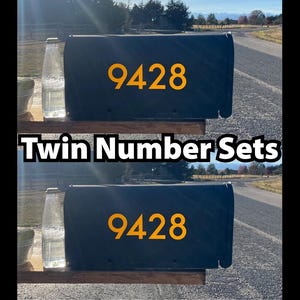 May include: Two black mailboxes with the number "9428" in gold. The text "Twin Number Sets" is displayed below the mailboxes. The mailboxes are on a wooden surface, with a bottle on the left side.