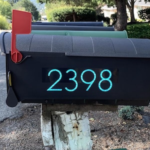 Op de afbeelding: Een zwarte brievenbus met het nummer 2398 in een turquoise lettertype. De brievenbus heeft een rode vlag en is gemonteerd op een houten paal. Andere brievenbussen zijn zichtbaar op de achtergrond.