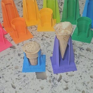 May include: A collection of colorful ice cream cone holders in various shades, including red, pink, orange, yellow, green, blue, and purple. Two holders are holding ice cream cones. The holders are made of plastic and are designed to prevent drips.