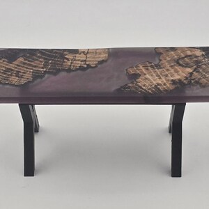 May include: A rectangular bench featuring a live-edge wood slab top with a translucent purple resin inlay. The wood displays a natural grain pattern with dark, textured edges. The bench is supported by four black metal legs angled outward.