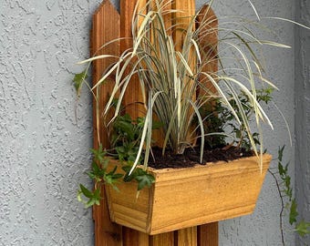 May include: A wooden wall-mounted planter box with a natural finish, filled with green and white plants. The planter is attached to a decorative wooden fence-like backing. The background is a textured grey wall.