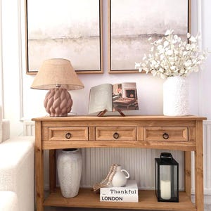May include: A wooden console table with three drawers and a lower shelf. It features a lamp, artwork, a vase of white flowers, and decorative items. The table has a natural wood finish, creating a warm and inviting aesthetic.