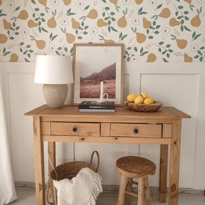 May include: A wooden console table with two drawers, a lamp, and a framed picture. A wooden bowl of lemons sits on the table. A woven basket with a white blanket and a small wooden stool are in front of the table. The wall has a pear pattern.