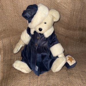 Pode incluir: Um urso de pelúcia vestindo um casaco de veludo azul marinho com botões dourados, um chapéu combinando e acabamento em pele branca. O urso tem um nariz preto e segura uma pequena bolsa azul marinho com uma estrela dourada.