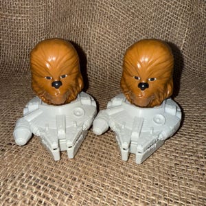 Op de afbeelding: Twee Chewbacca-beeldjes met bruine vachtkoppen en zwarte neuzen zitten op grijze Millennium Falcon-modellen. Het speelgoed is gemaakt van plastic en is waarschijnlijk verzamelobjecten of kinderspeelgoed. De achtergrond is een bruin oppervlak.