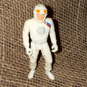 May include: A vintage white action figure of a space pilot or astronaut. The figure is wearing a white helmet with orange goggles, a white jumpsuit, and white boots. The figure has a gray belt and a patch with red and blue stripes on the shoulder.
