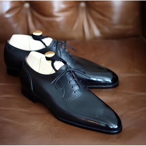 Pu&ograve; includere: Un paio di scarpe eleganti in pelle nera con finitura lucida, caratterizzate da un classico design stringato e perforazioni decorative. Le scarpe sono esposte su una superficie in pelle marrone, con forme di legno all'interno.